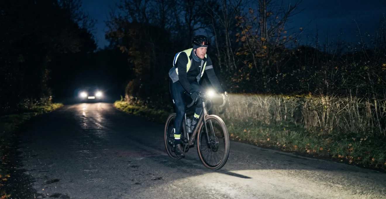 A cyclist rides on an unlit rural road at night with a front bike light aimed low on the asphalt while a car approaches in the distance.